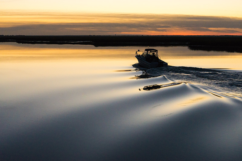 boat-at-sunset Boat cruising across calm water at sunset, leaving ripples behind with a golden sky in the background.