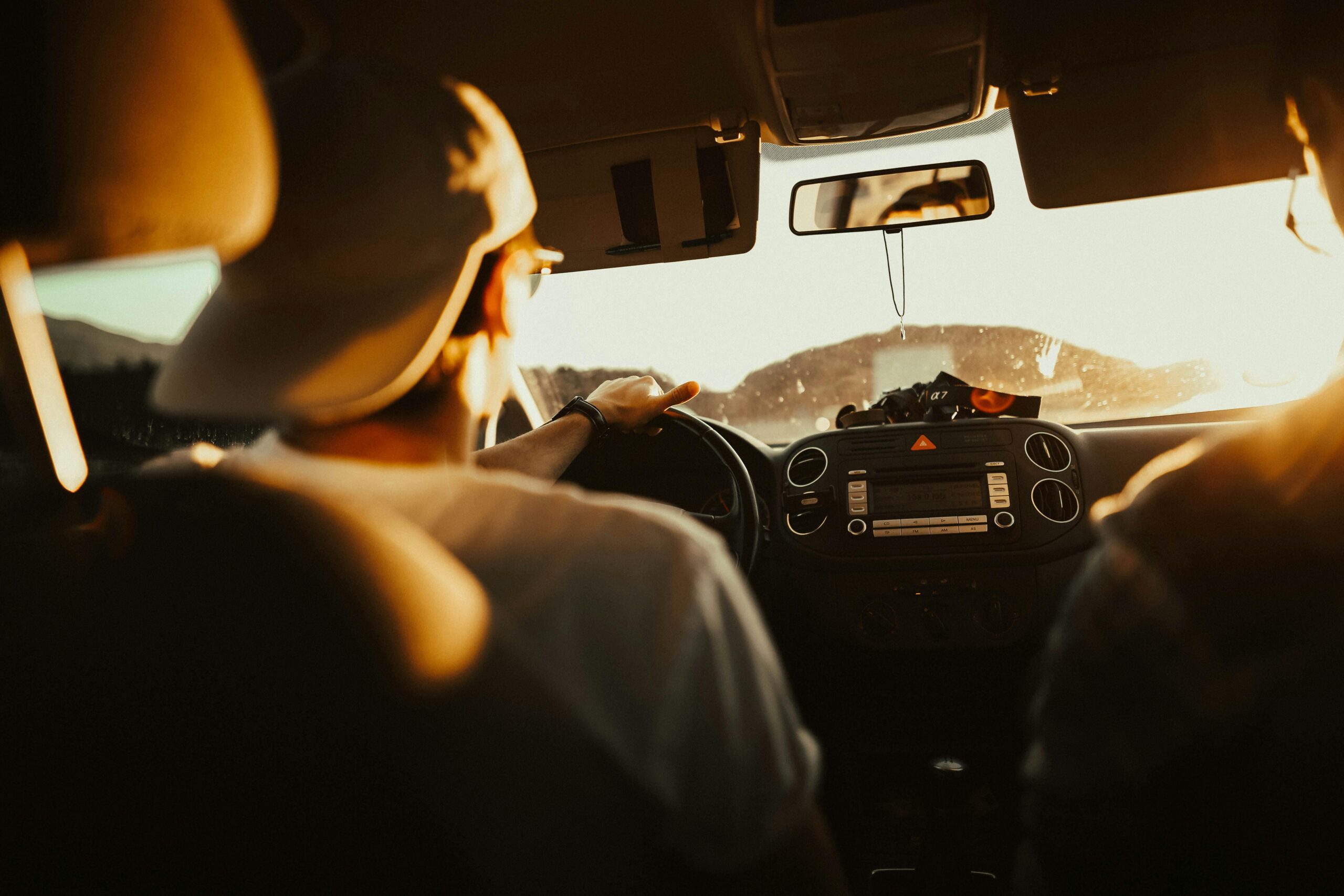 pexels-pripicart-620335 Driver wearing a cap steering a car at sunset with warm sunlight shining through the windshield.